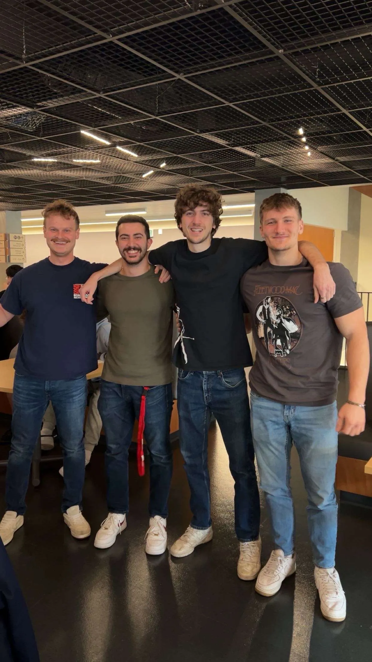 Four friends standing arm in arm indoors under a black grid ceiling, all wearing jeans and white sneakers.