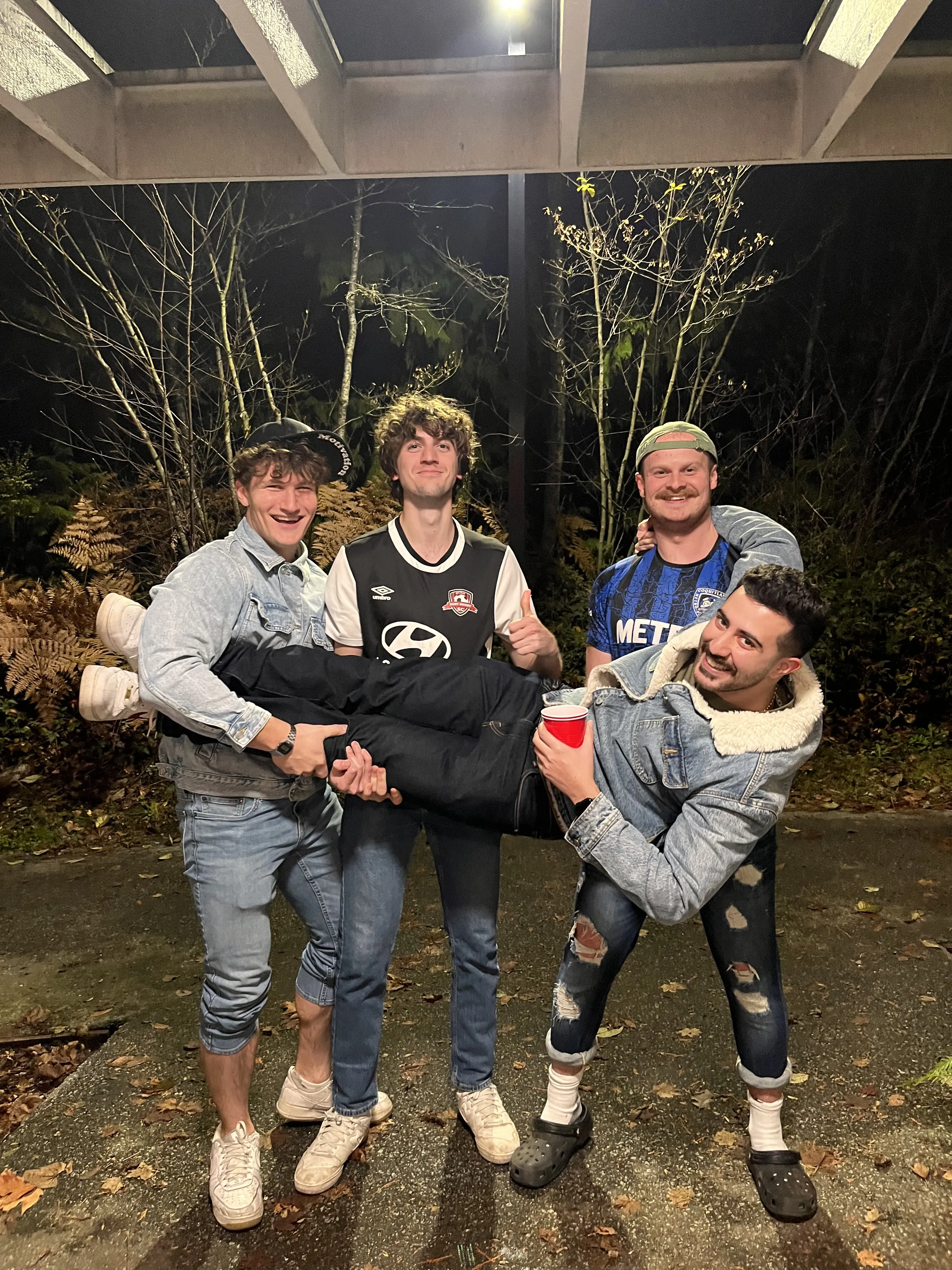 Five friends outside at night under a covered walkway, in denim jackets and jeans. Two of them carry a third sideways across their arms while the other two pose behind, one giving a thumbs up.